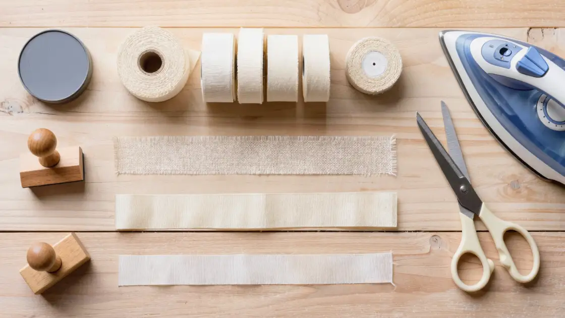 Rollos de cinta de algodón, sello, tinta textil, tijeras y plancha sobre una mesa de madera